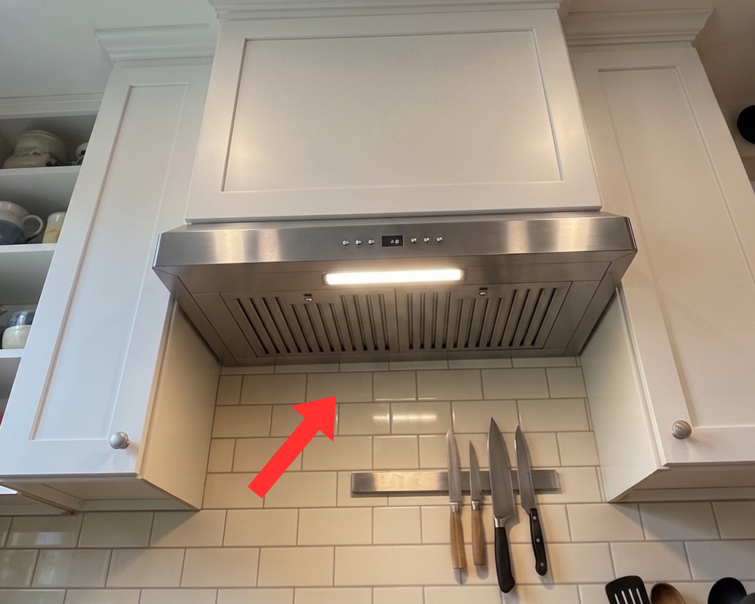 Stainless steel ducted range hood with its light on, built into white cabinetry above a subway tile backsplash
